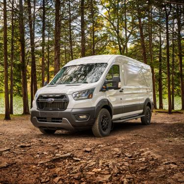 Photo of white Ford Transit Trail van from front 3/4 view, with black trim and offroad tires, on hard packed dirt with tall pine trees and water behind it.